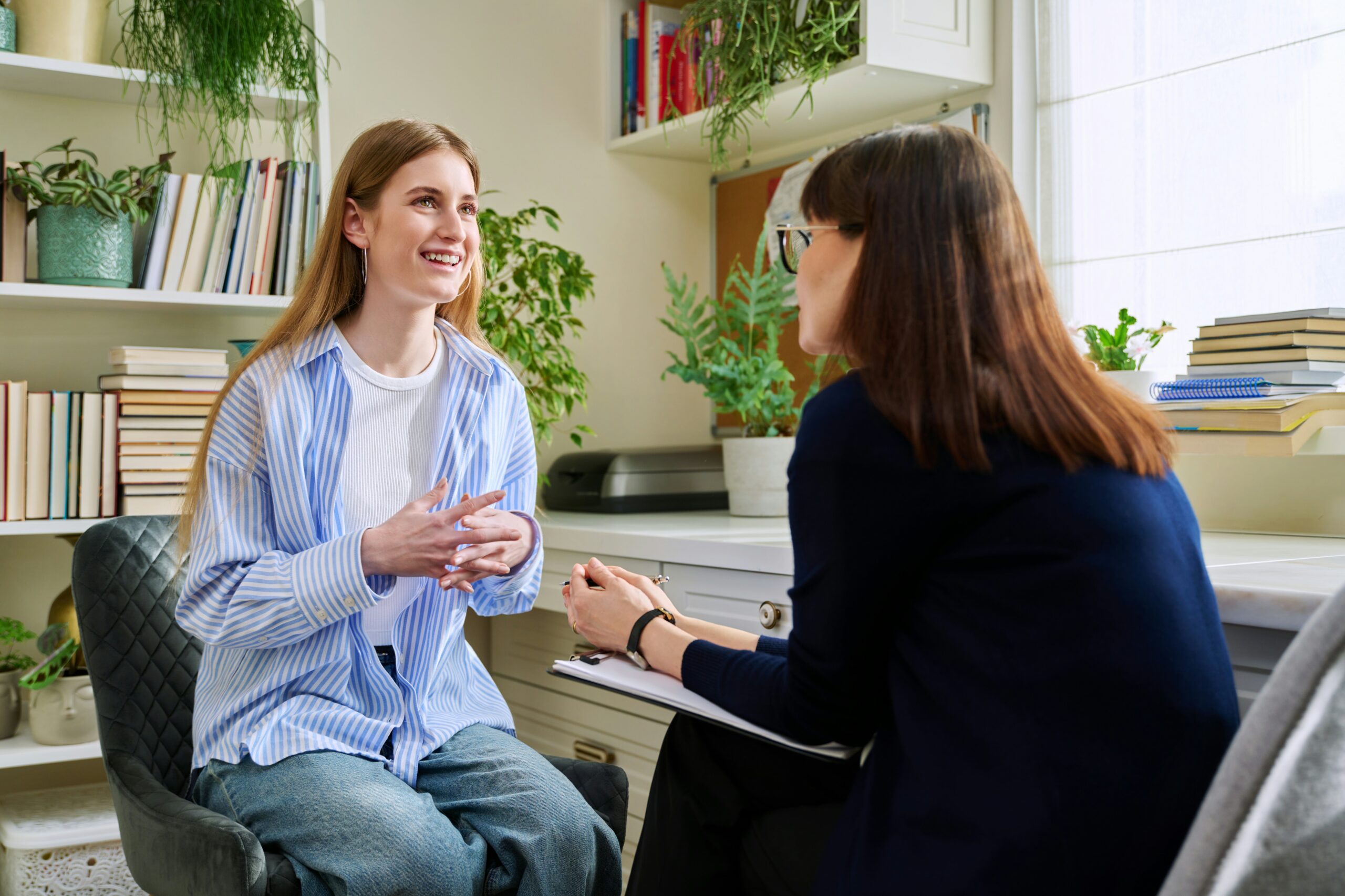 A photograph of a smiling young woman gesturing with her hands while speaking to a counselor in an office setting. The counselor sits opposite her holding a clipboard, listening attentively in a bright room filled with books and potted plants.
