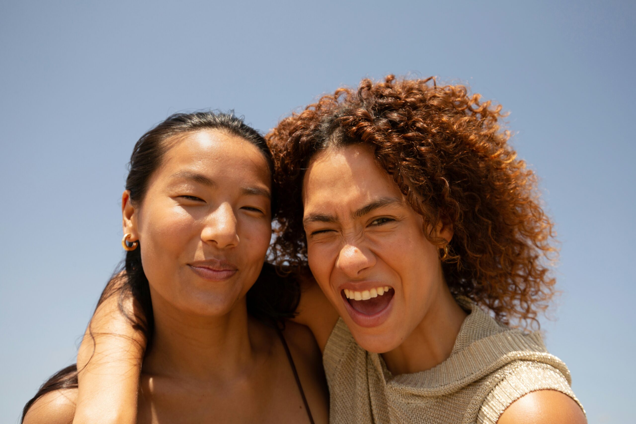 A close-up photograph of two smiling friends posing close together against a clear blue sky background. The woman on the left smiles gently at the camera, while the woman on the right with curly brown hair laughs enthusiastically with her arm around her friend's shoulder.