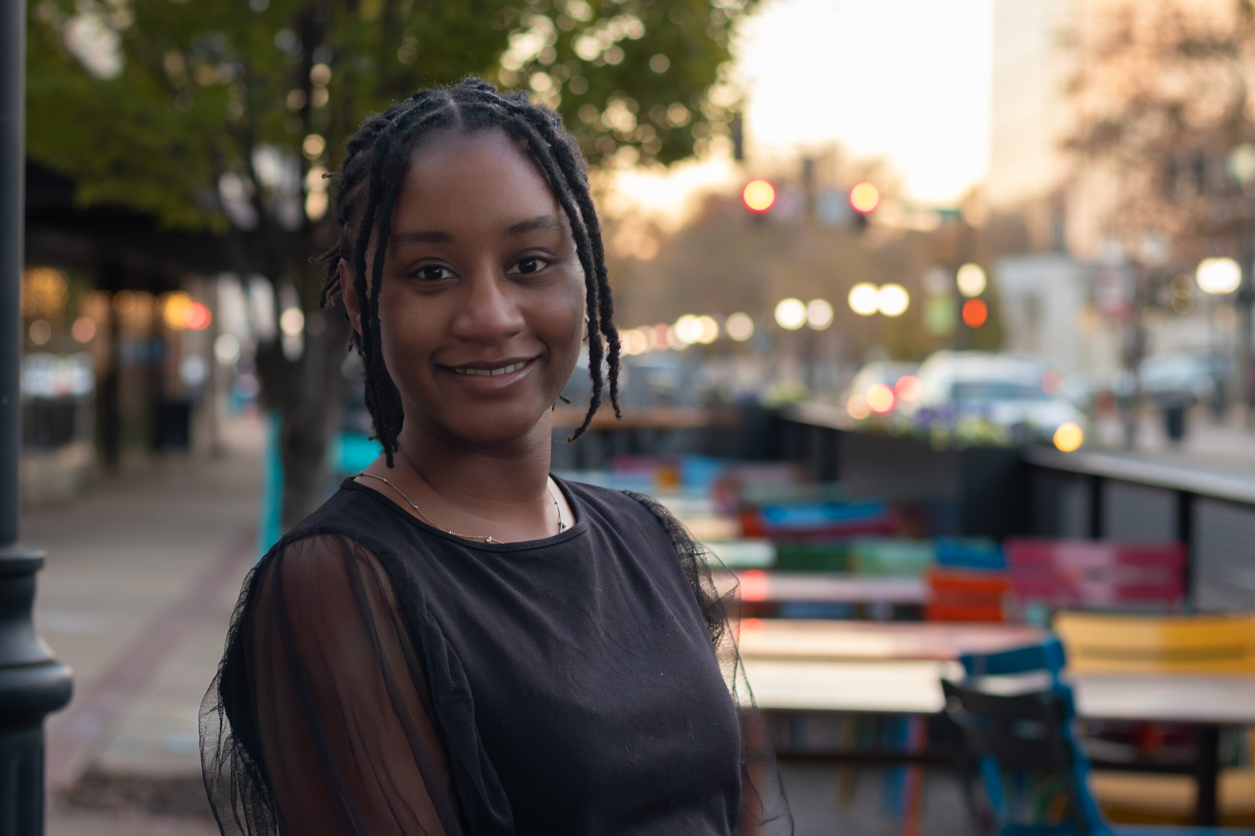 A photograph of a Chakiah with braided hair smiling gently at the camera. She stands outdoors next to an empty open-air patio with colorful chairs and tables, with a softly blurred city street visible in the background during dusk.