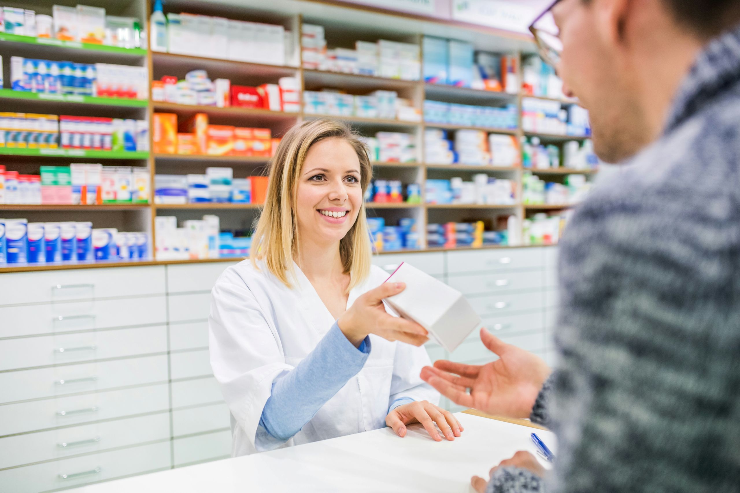 A smiling female pharmacist in a white lab coat hands a small, white medicine box to a customer over the counter. The background shows well-organized pharmacy shelves stocked with various medication packages.
