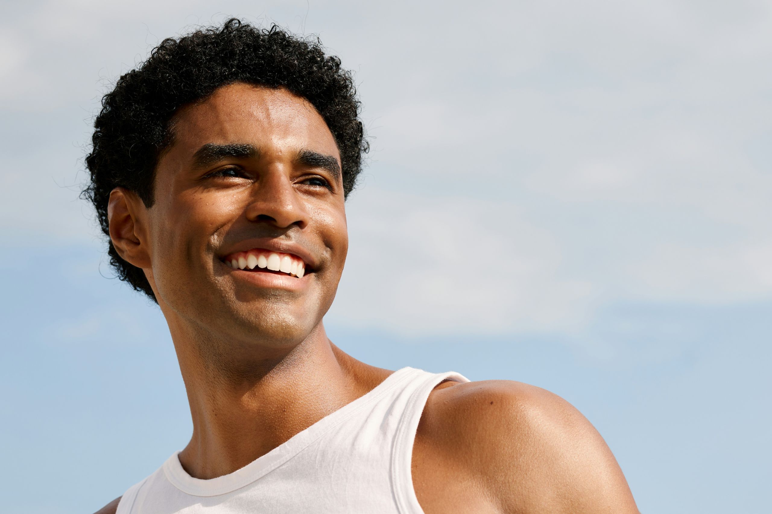A low-angle portrait of a young man with short, curly black hair wearing a white tank top and smiling broadly. He is looking off to the side against a clear, bright blue sky background.