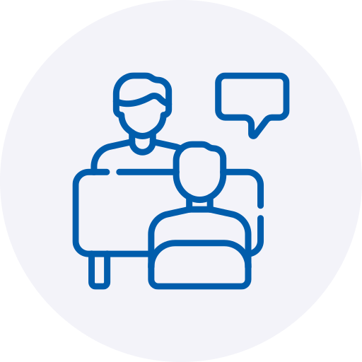 A blue line-art icon depicting two people in a meeting or therapy session, with one individual sitting behind a desk or table and the other seated across from them. A blank speech bubble hovers near the person behind the desk, all set against a soft, light grey circular background.