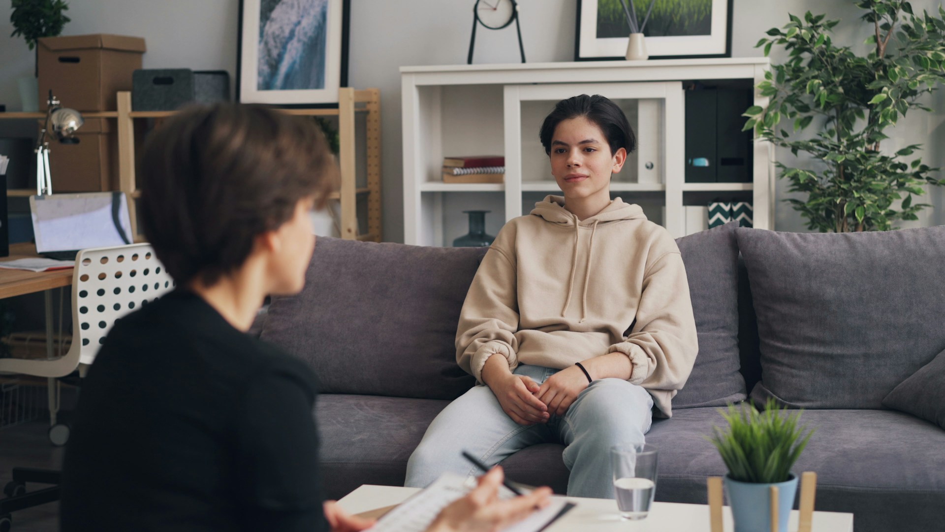 A photograph of a young person wearing a beige hoodie sitting on a grey couch during a therapy or counseling session. They listen calmly while looking towards a therapist who sits in the foreground with their back to the camera, holding a clipboard and pen.
