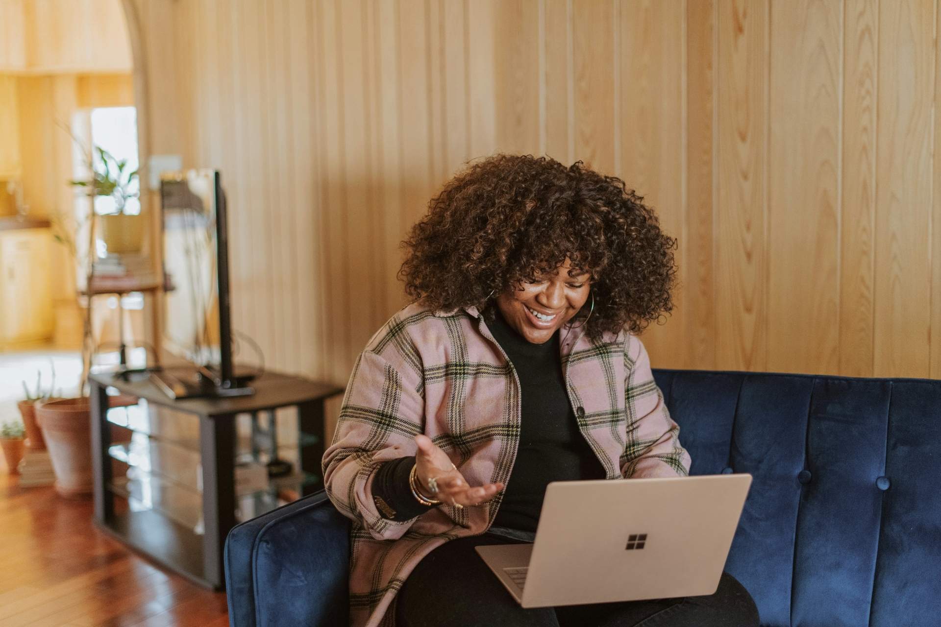 A photograph of a smiling woman with voluminous curly hair sitting on a blue velvet sofa while using a laptop. She gestures with one hand toward the screen, set within a cozy room featuring light-colored wood paneled walls.