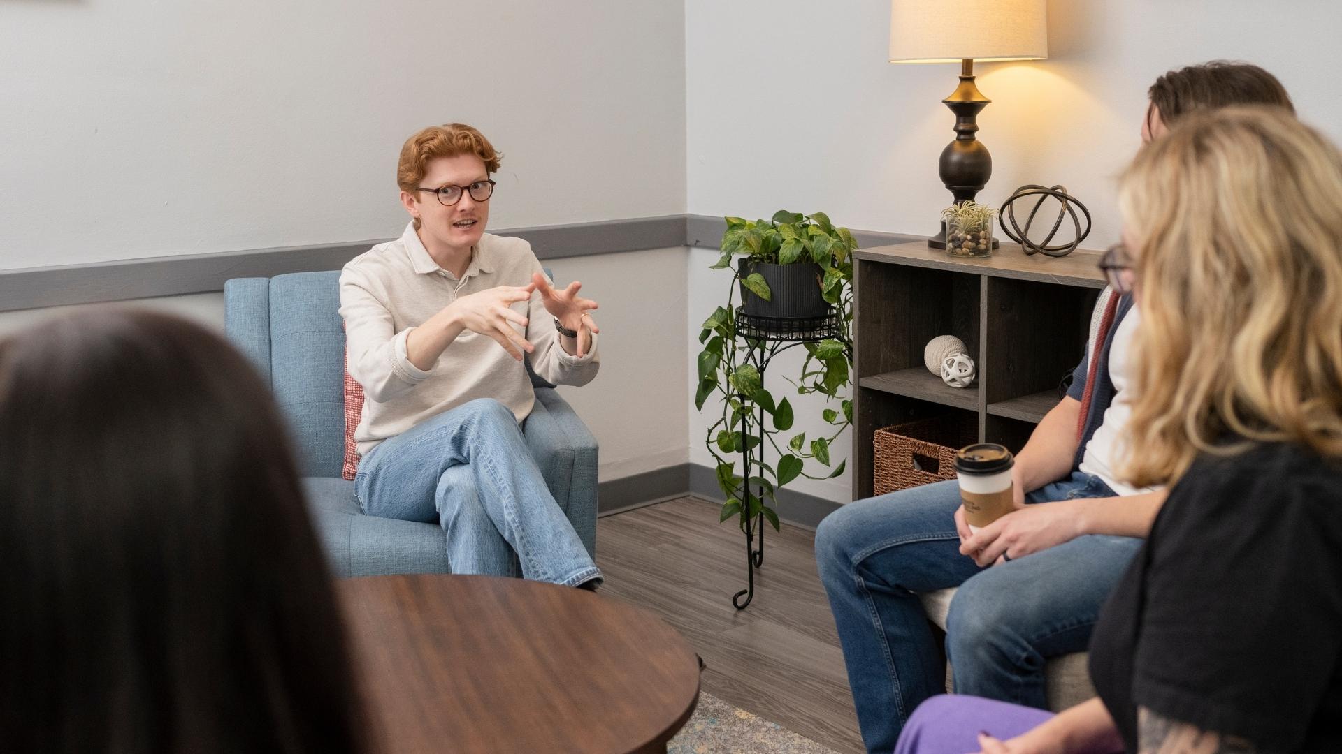 A small group participates in a counseling or support session in a comfortable office setting, seated in a circle and engaged in open discussion. The welcoming environment encourages connection, communication, and personal growth through shared experiences and guided conversation.