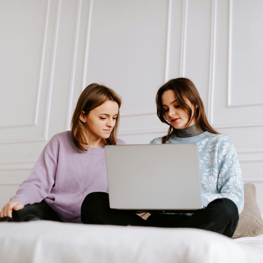 Two young women sitting on a bed, looking at a laptop together. They are indoors in a bright room with white paneled walls, wearing casual sweaters, and appear focused on the screen.