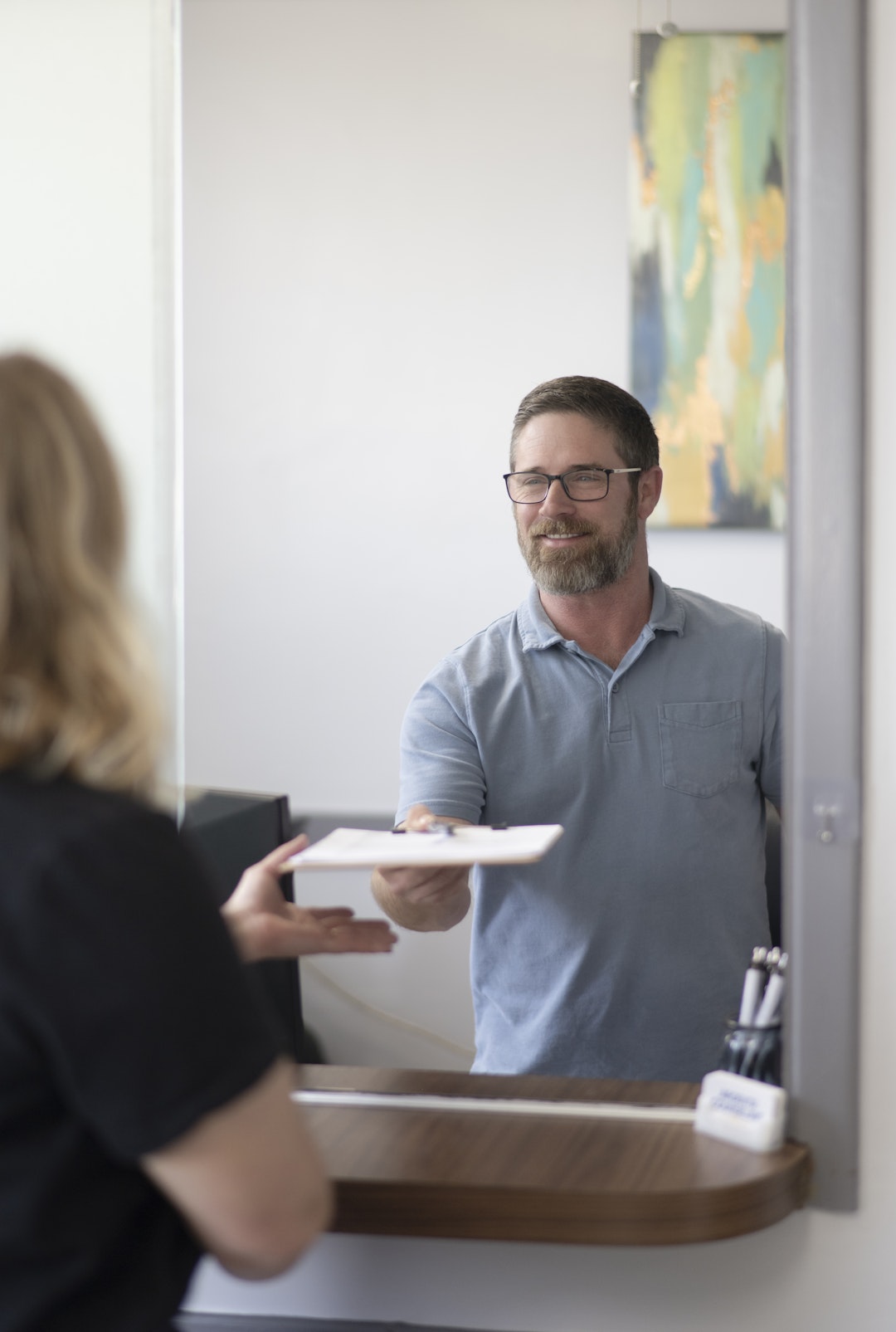 A staff member hands paperwork to a visitor at a reception desk, creating a welcoming and professional first impression. The image highlights friendly customer service and a smooth check-in experience in a counseling and wellness office.