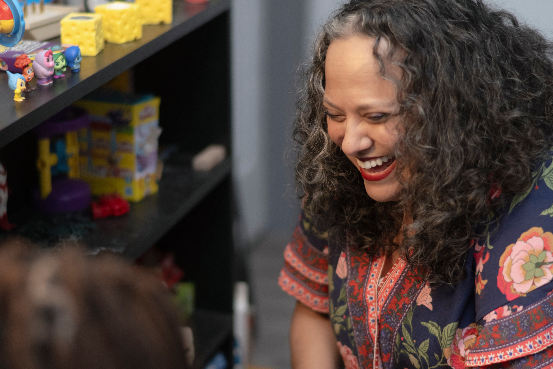 Counselor smiling and engaging with a client in a welcoming therapy space, seated near a shelf of toys, games, and therapeutic tools. The warm interaction creates a supportive environment for child or family counseling.