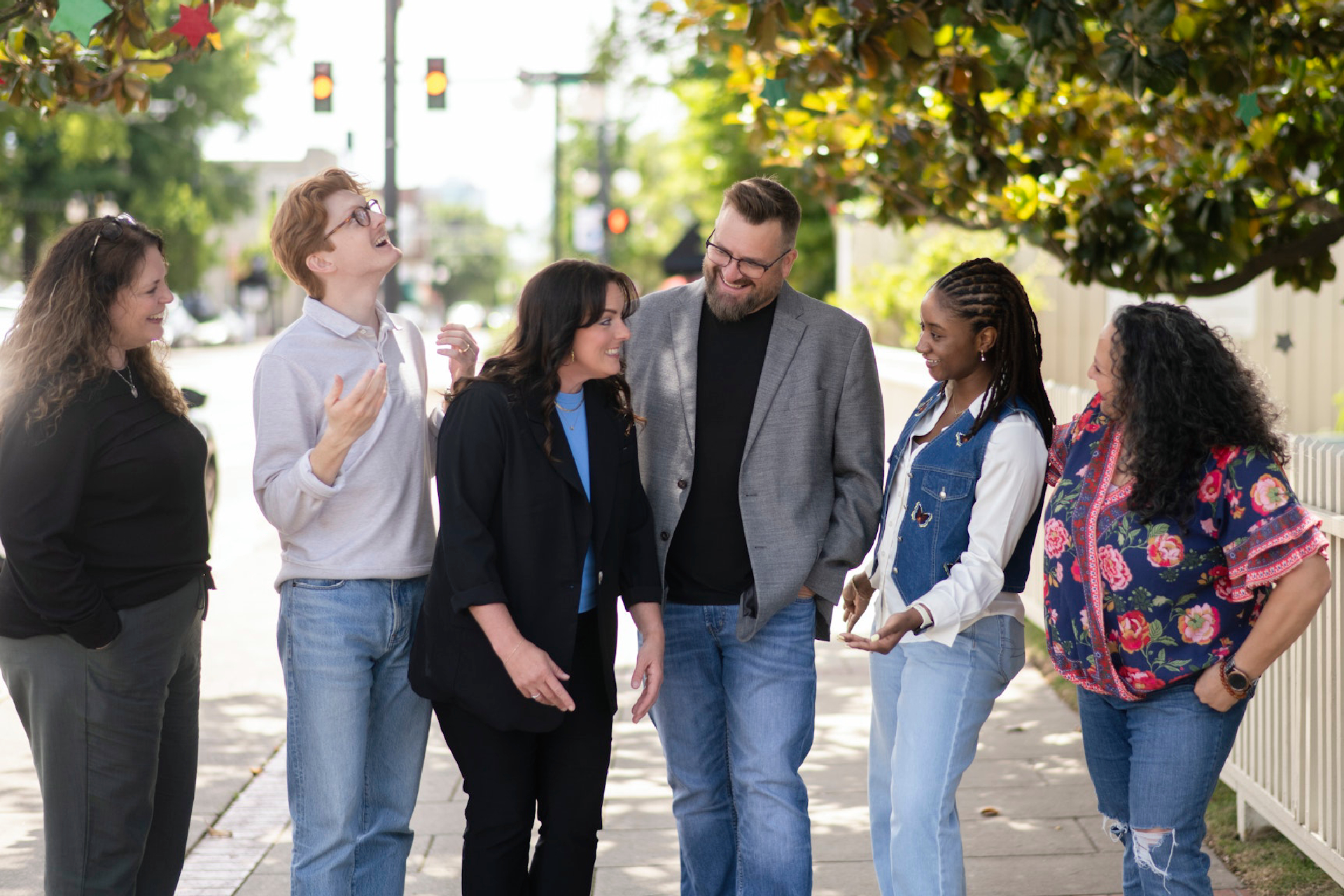 LSP_3152 (1) Group of six adults standing outdoors on a sidewalk, smiling and talking together in a relaxed and supportive atmosphere. The diverse group appears engaged in friendly conversation beneath leafy trees in a bright neighborhood setting.
