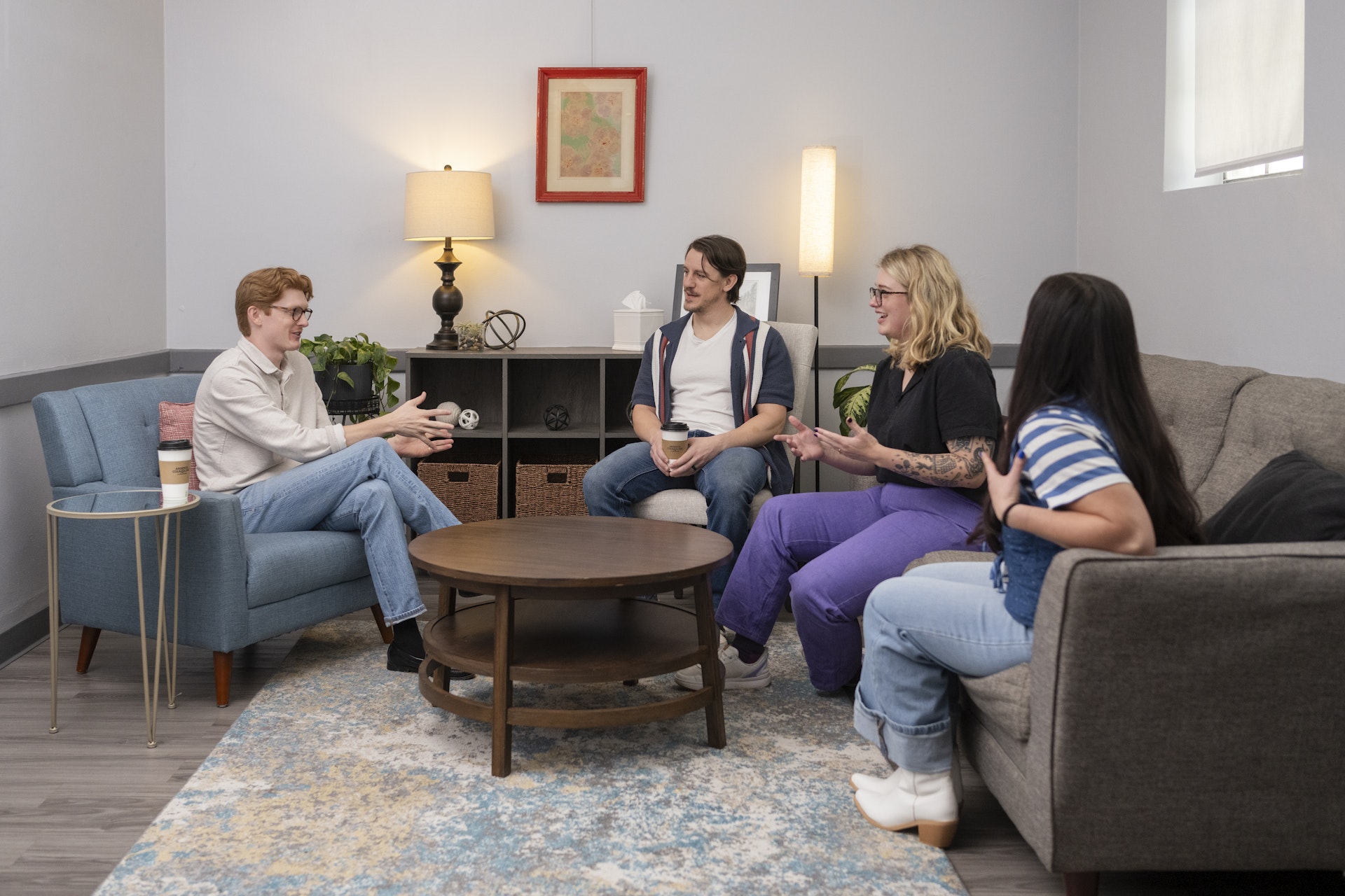 A small group participates in a counseling or support session in a comfortable office setting, seated in a circle and engaged in open discussion. The welcoming environment encourages connection, communication, and personal growth through shared experiences and guided conversation.