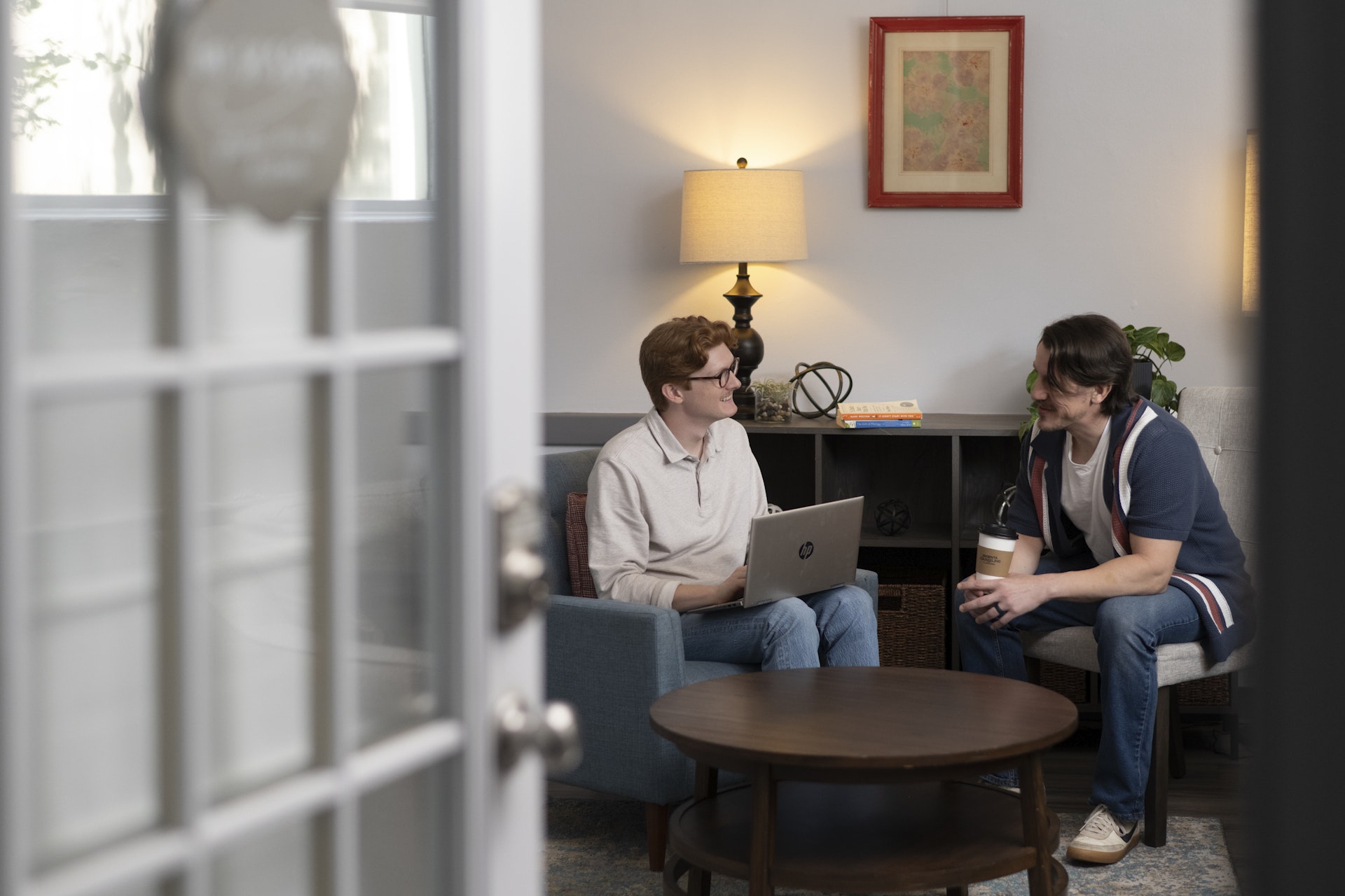 Two people sit in a comfortable counseling office, engaged in a friendly conversation while one uses a laptop. The warm lighting, cozy furnishings, and open office setting create an inviting environment for support, collaboration, and meaningful discussion.