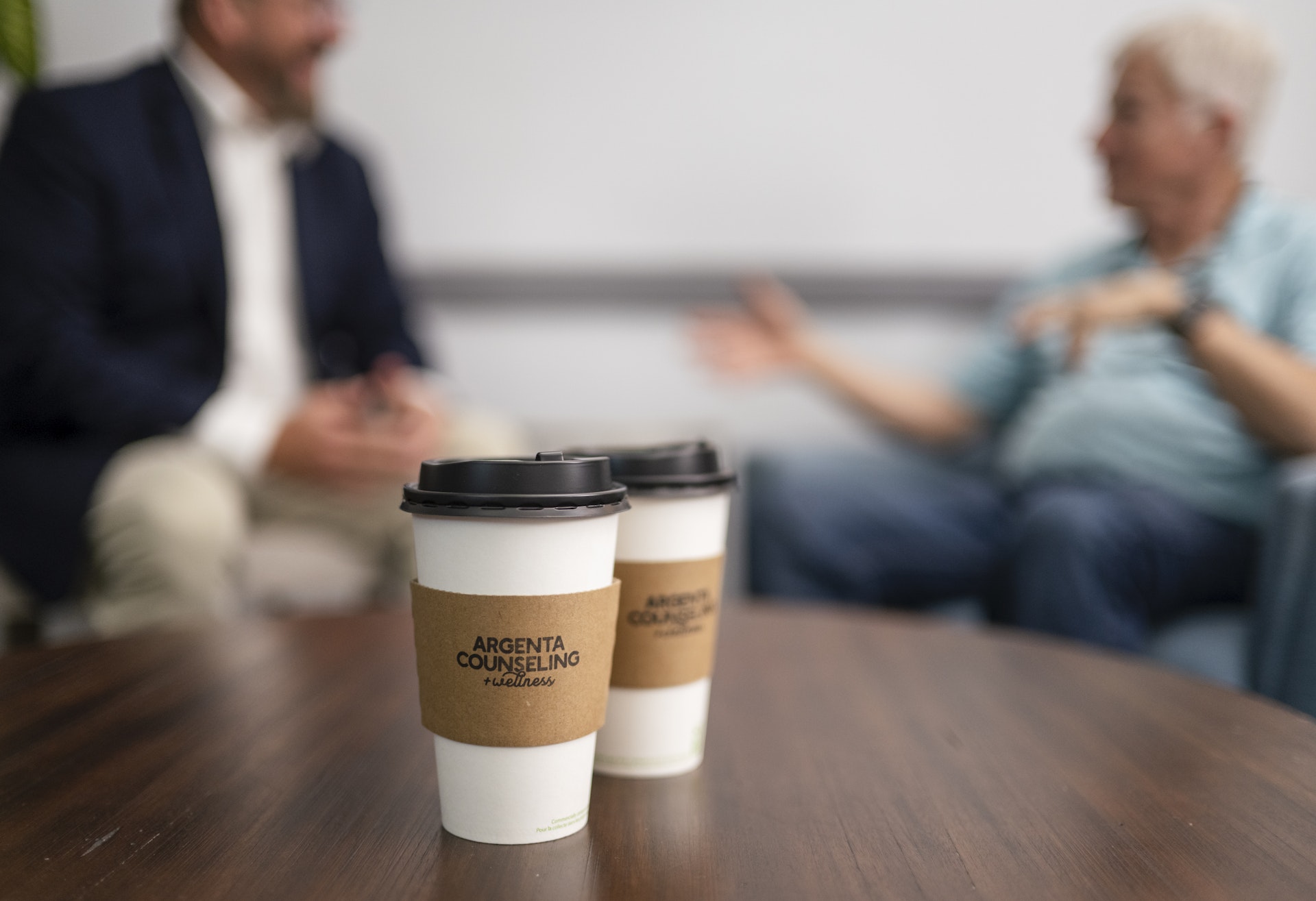 LSP_3333 Two branded coffee cups sit on a table in the foreground while a counselor and client engage in conversation in a comfortable office setting. The blurred background highlights a warm, welcoming environment focused on connection, support, and wellness.