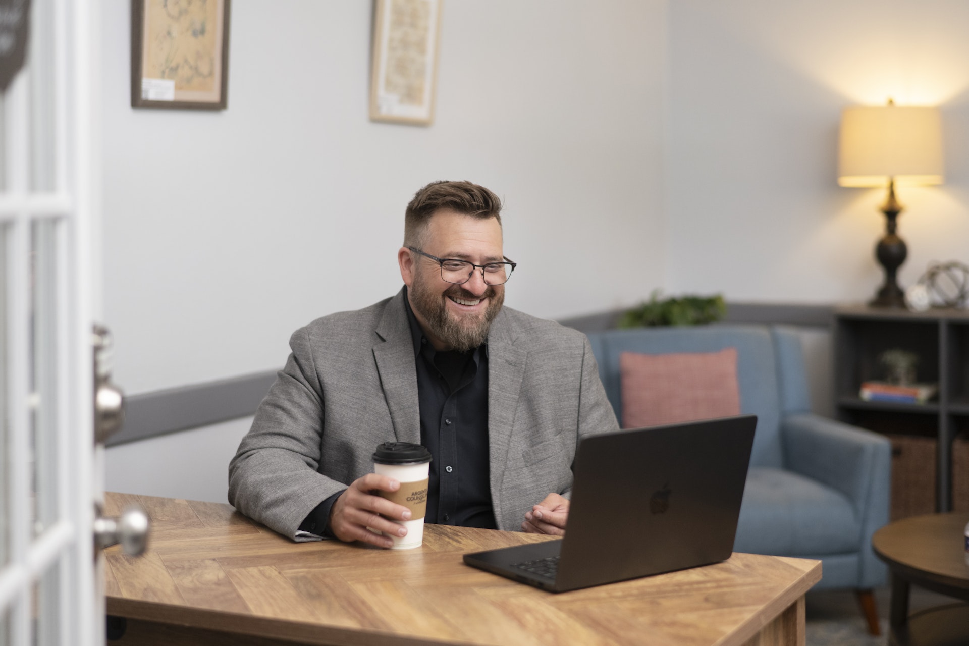 Man smiling during a virtual counseling or coaching session, seated at a desk with a laptop and a takeaway coffee cup in a comfortable office setting. He appears engaged and approachable while participating in an online meeting.