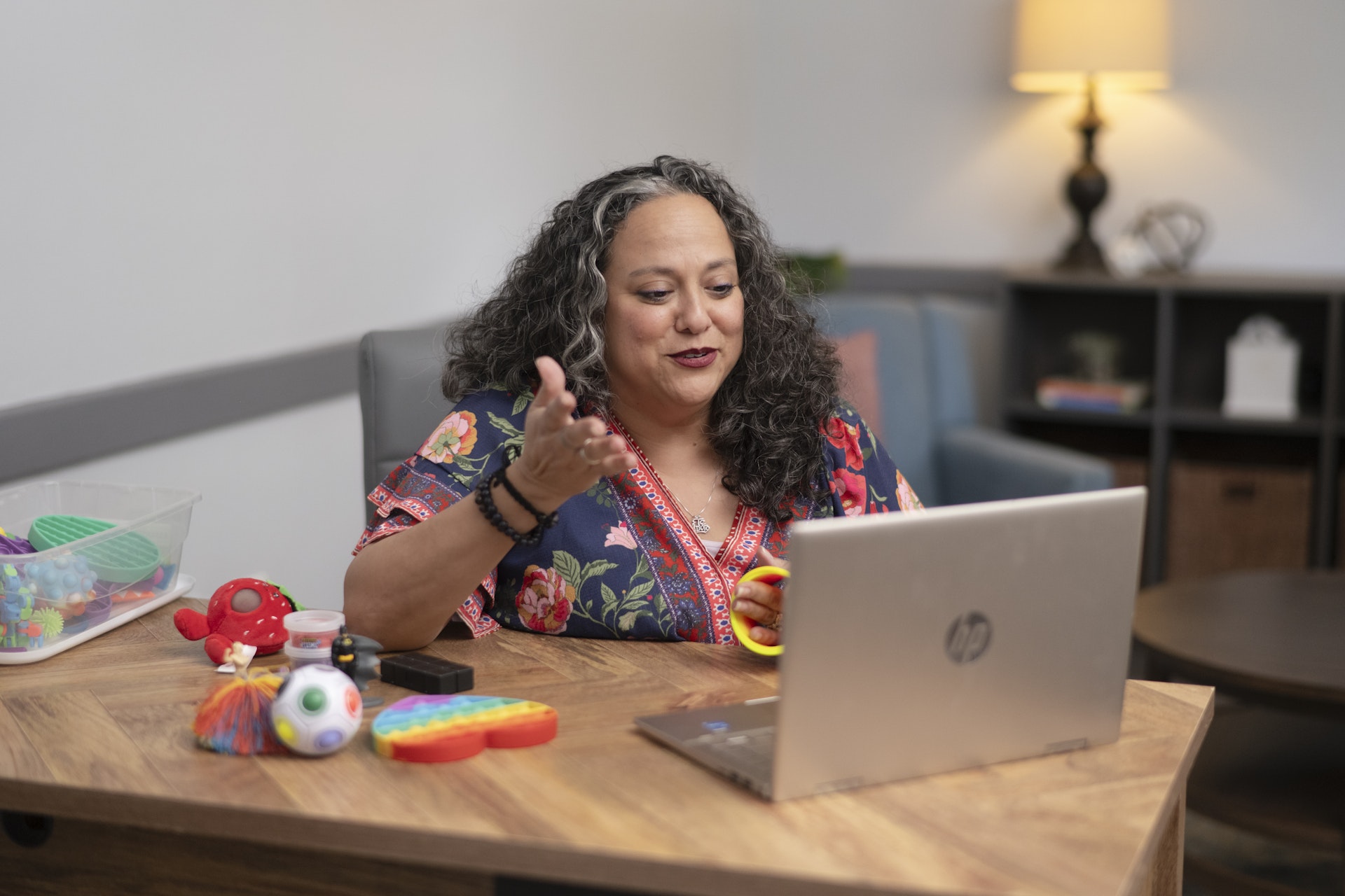 Woman participating in an online counseling or therapy session while seated at a desk with a laptop. She gestures as she speaks and uses sensory tools on the table, creating an engaging and supportive virtual counseling environment.