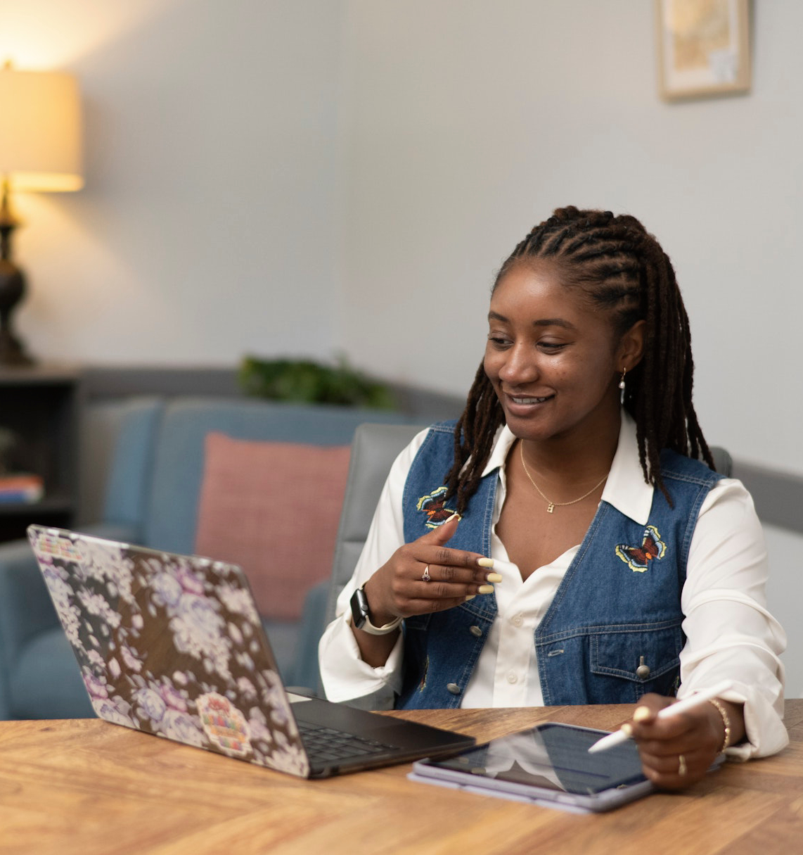 LSP_3481 (1) A woman sits at a table in a comfortable office, using a laptop and tablet while participating in a virtual meeting or telehealth session. The warm, welcoming environment highlights modern, flexible counseling and professional support services.