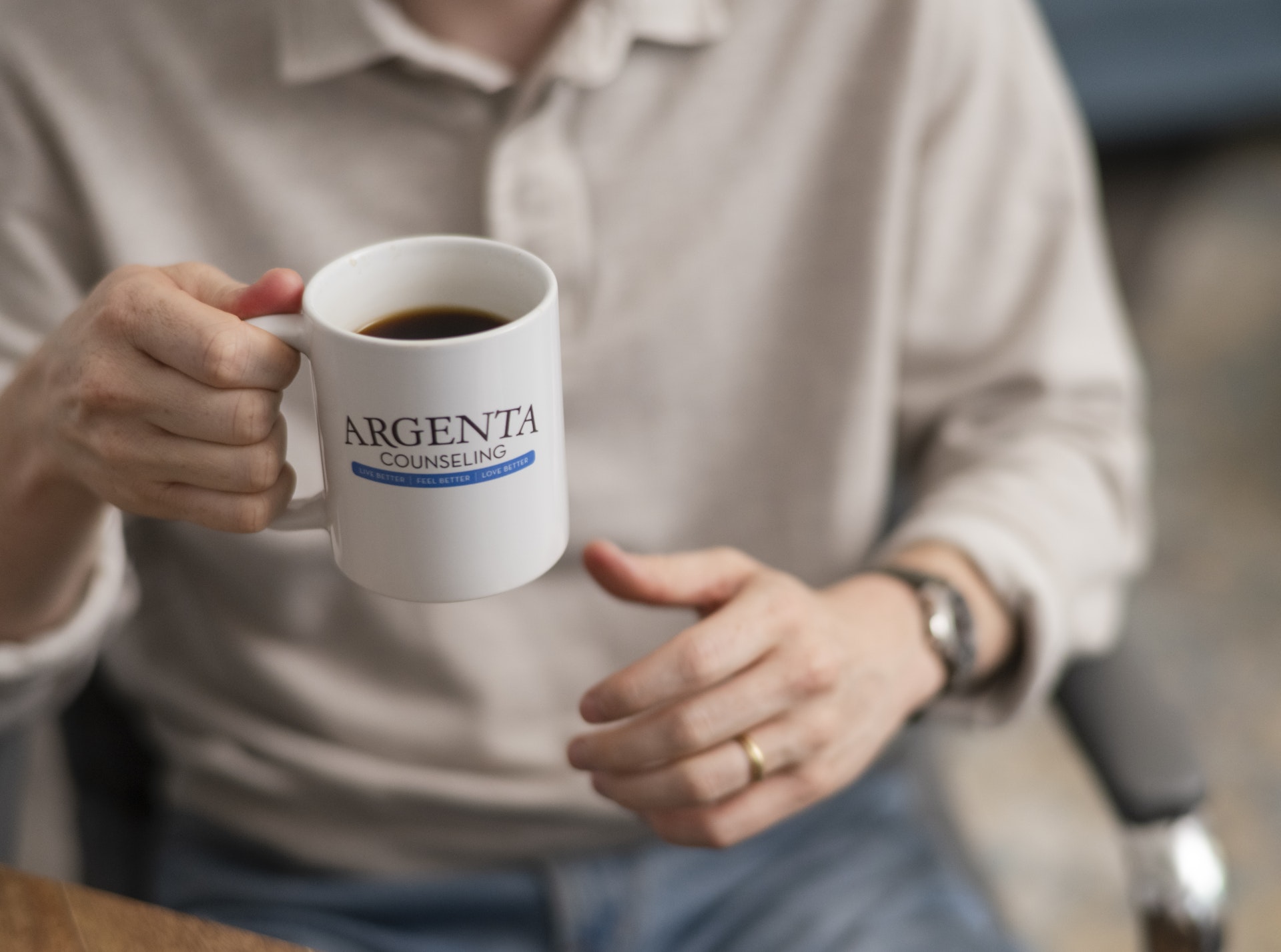 A person holds a branded coffee mug while seated in a comfortable office setting, creating a warm and inviting atmosphere. The image conveys a sense of connection, comfort, and support often associated with counseling and wellness services.