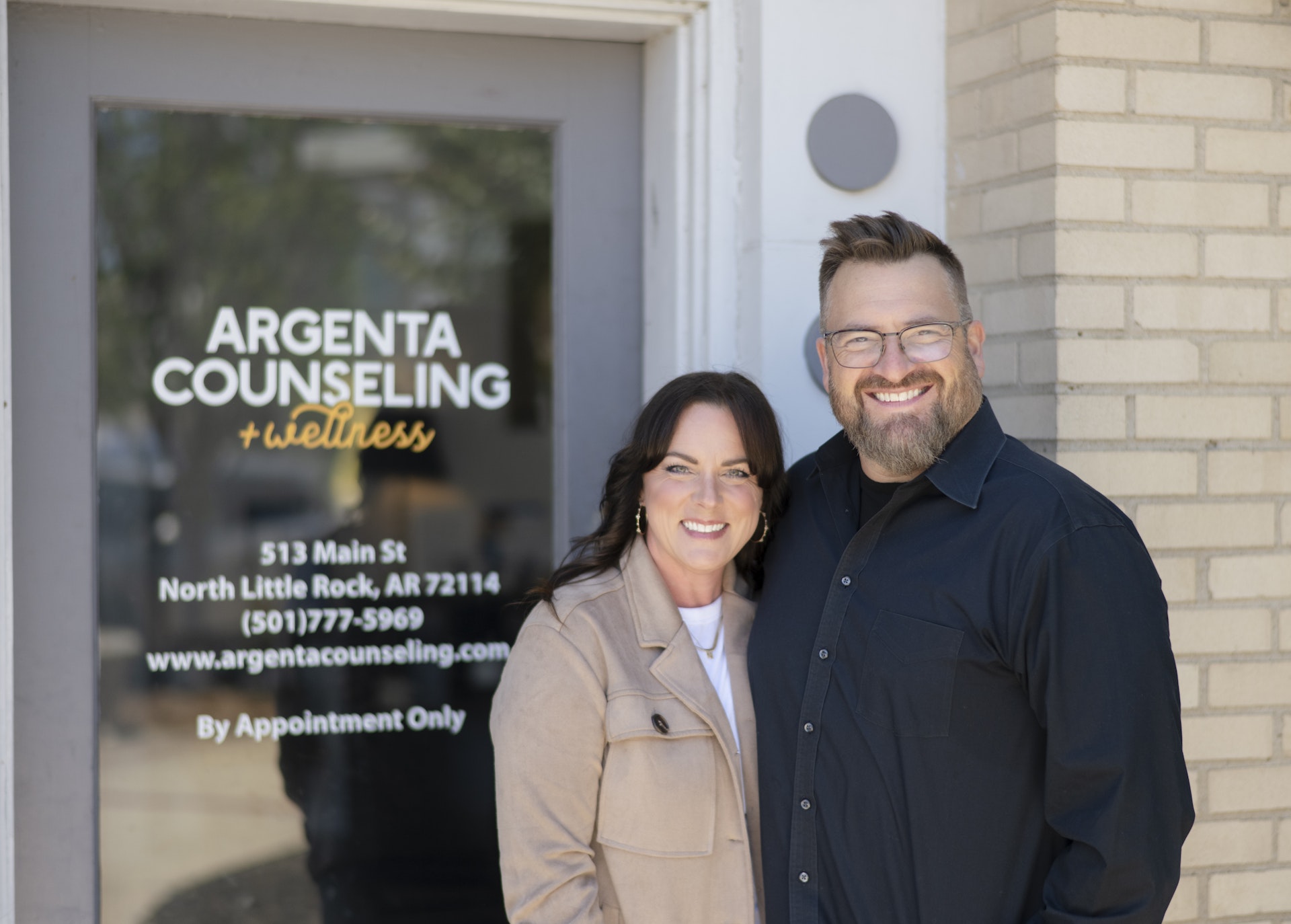 Two professionals stand outside the entrance of a counseling and wellness practice, smiling in front of the office door. The image highlights a welcoming, approachable team dedicated to providing compassionate counseling and mental health services.
