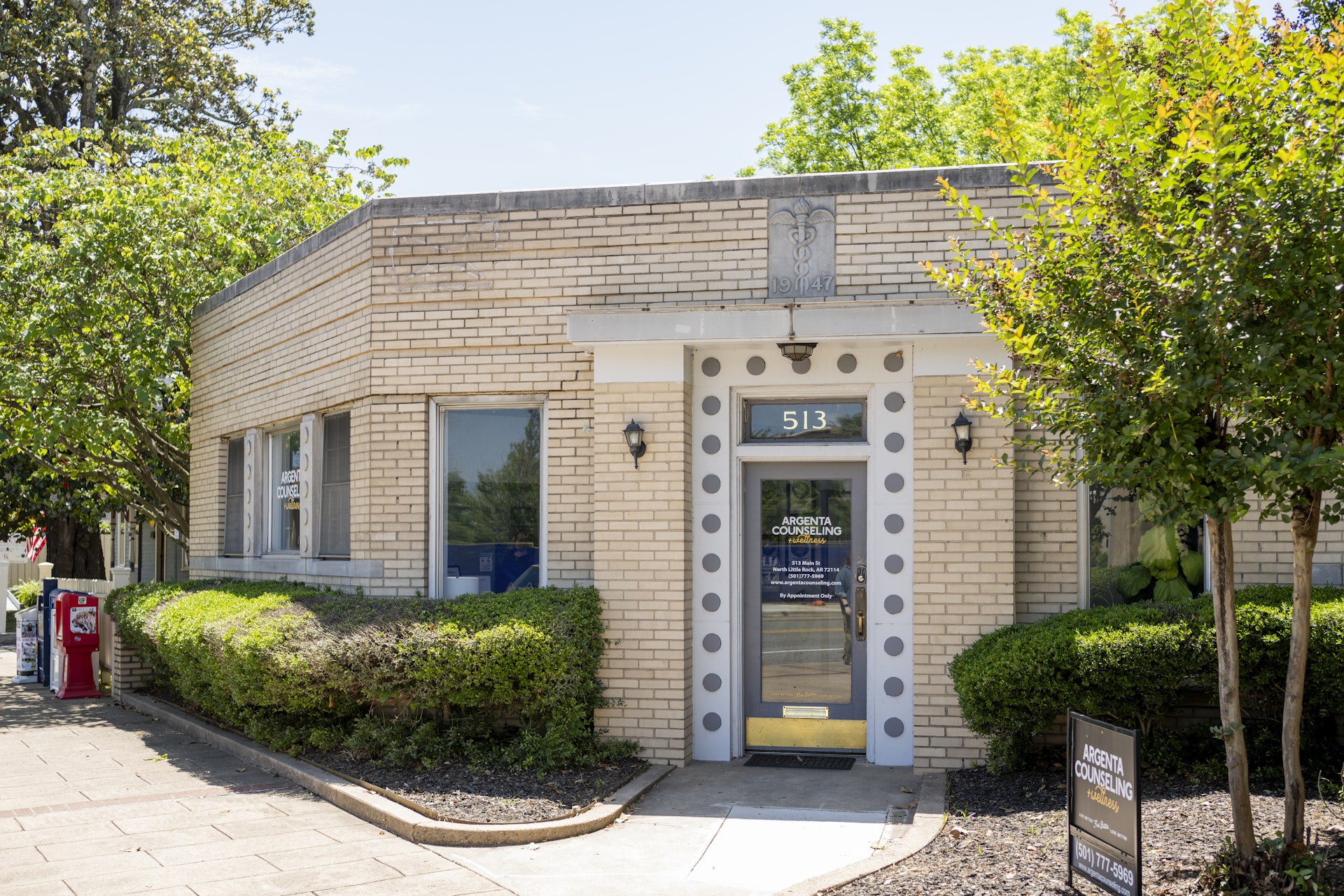 Exterior view of a counseling office building with landscaped shrubs, mature trees, and a clearly marked entrance along a sidewalk. The welcoming professional office is located in a standalone brick building with signage visible near the front entrance.