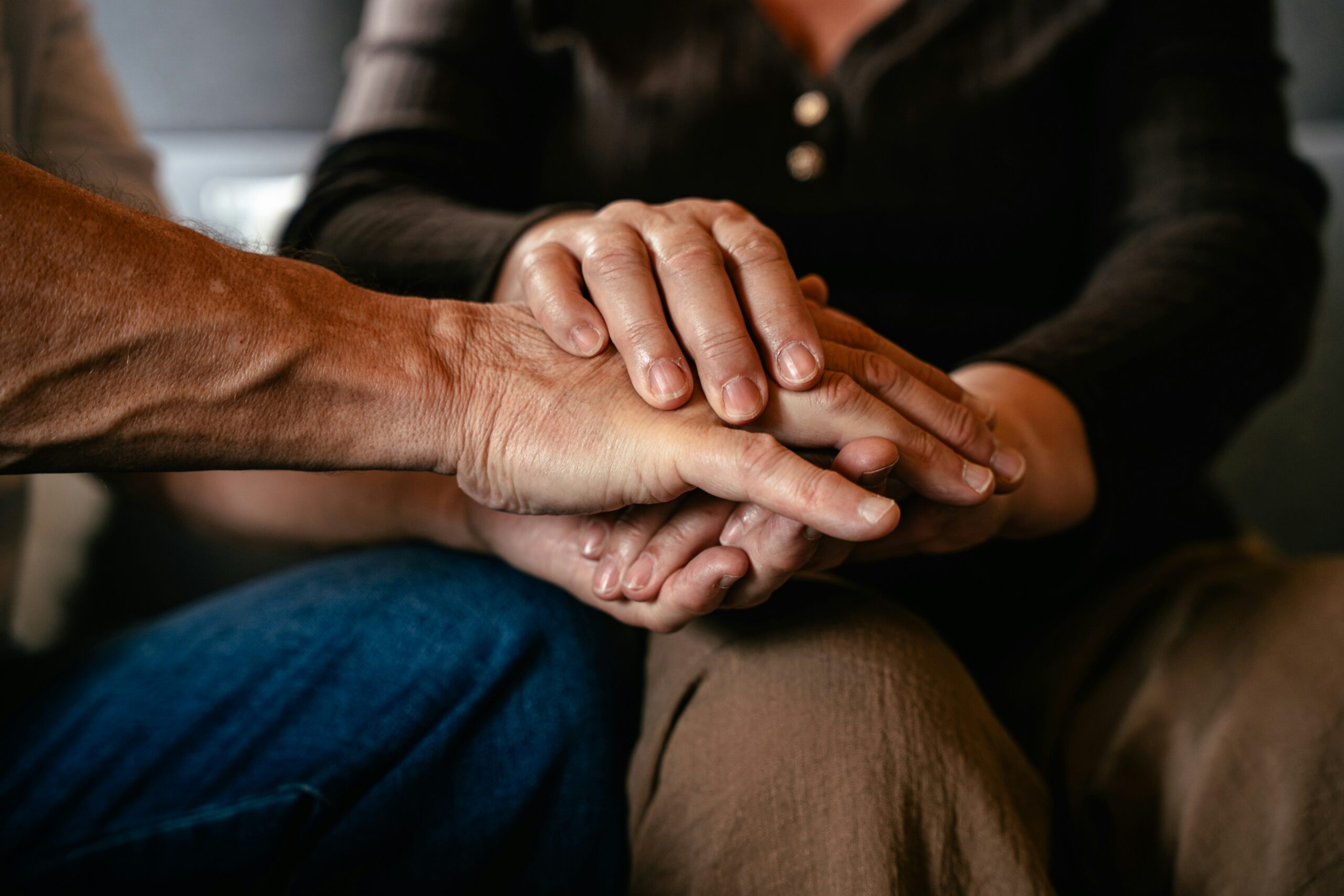 A close-up photograph of three hands gently stacked on top of one another, symbolizing comfort, support, or solidarity. The hands, showing signs of age, rest closely together in a dimly lit setting.