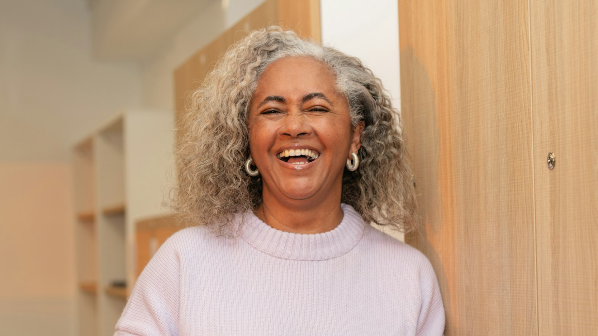 Smiling older woman with curly gray hair wearing a light lavender sweater and hoop earrings, standing indoors near a wooden door. She appears cheerful and approachable, laughing warmly in a bright, modern interior setting.