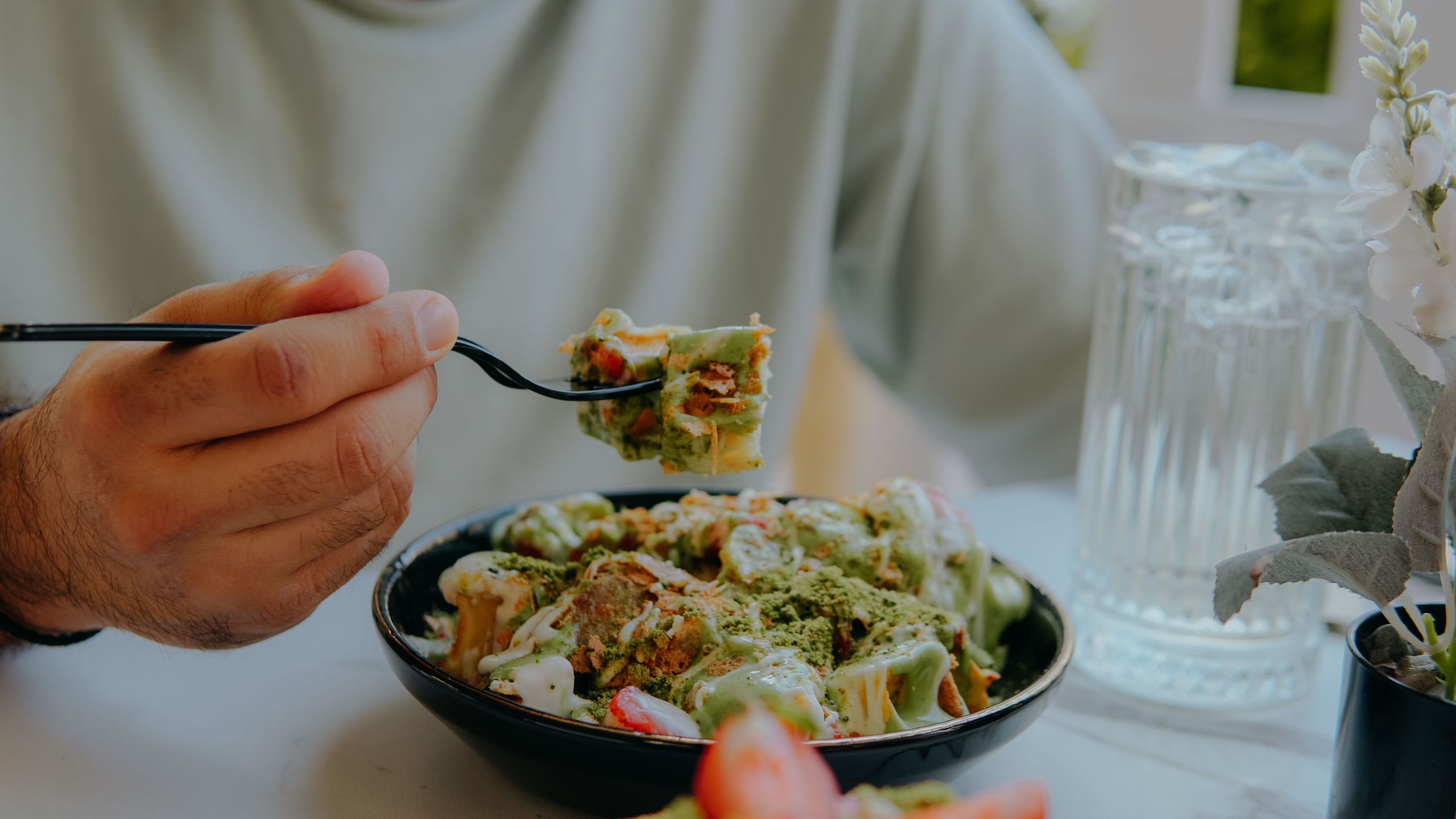 A close-up photograph of a person holding a fork with a bite of a dessert drizzled in green matcha sauce and white cream. The black bowl contains sliced strawberries and pastry pieces dusted with matcha powder, alongside a glass of iced water.