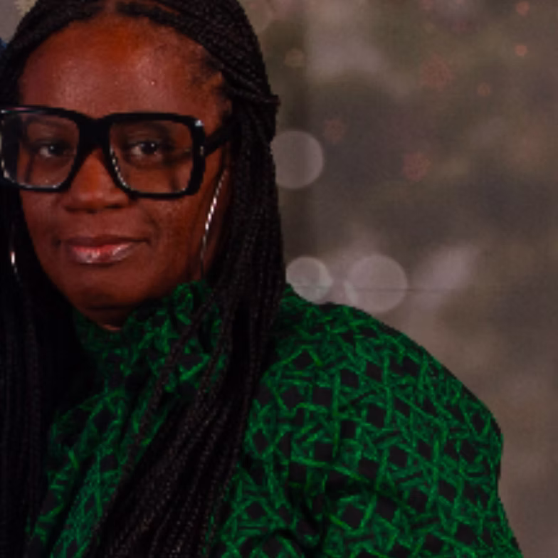 Kisha Floyd wearing black glasses and a green patterned top with long braids poses in front of a blurred background with circular light spots.