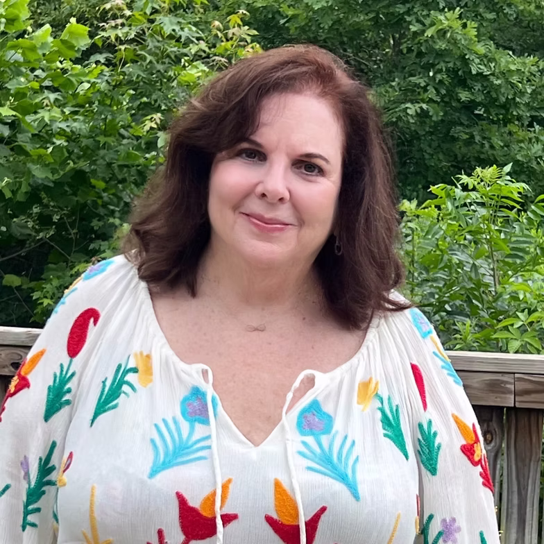 Group discussion-bro Ann Richardson Wrotny with shoulder-length brown hair, wearing a white blouse with colorful floral patterns, stands outdoors in front of green foliage and a wooden railing, smiling at the camera.