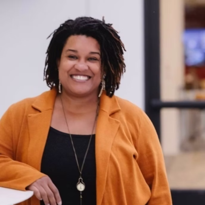 Shakira Eakins with medium-length curly hair, wearing a burnt orange blazer and black top, smiles while standing indoors with her hand resting on a white surface. The background is softly blurred.