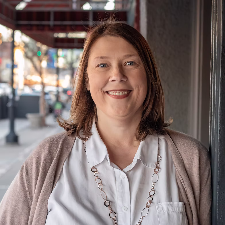 Anita Rutledge-Kuykendall with straight brown hair, wearing a white blouse, light cardigan, and necklace, smiles while standing outside on a city sidewalk with blurred buildings and lights in the background.