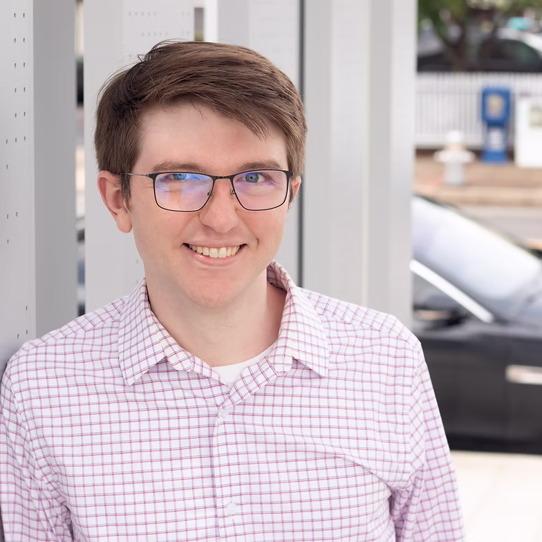 Nick Clemmons with short brown hair and glasses, wearing a white and pink plaid shirt, smiles while standing outdoors near a glass structure with a blurred car and street scene in the background.