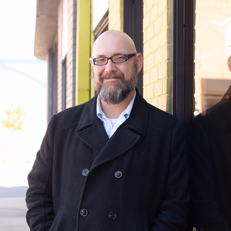 Jesse Hubberd wearing glasses and a black coat stands outside, leaning against a yellow brick wall and smiling at the camera.