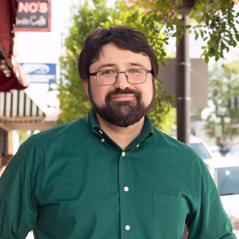 Michael Dillon with glasses and a beard, wearing a green button-up shirt, stands outdoors on a sunny day with trees and storefronts in the background.