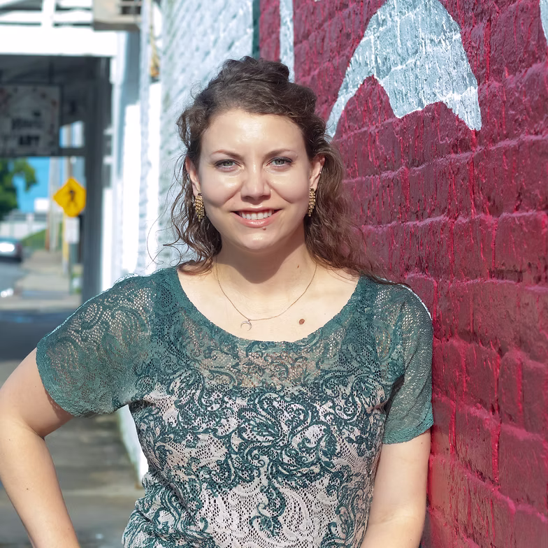Group discussion-bro Kate McCalman with curly hair smiles while standing against a red brick wall with white graffiti. She wears a green lace top and gold necklaces. The scene is outdoors on a sunny day.