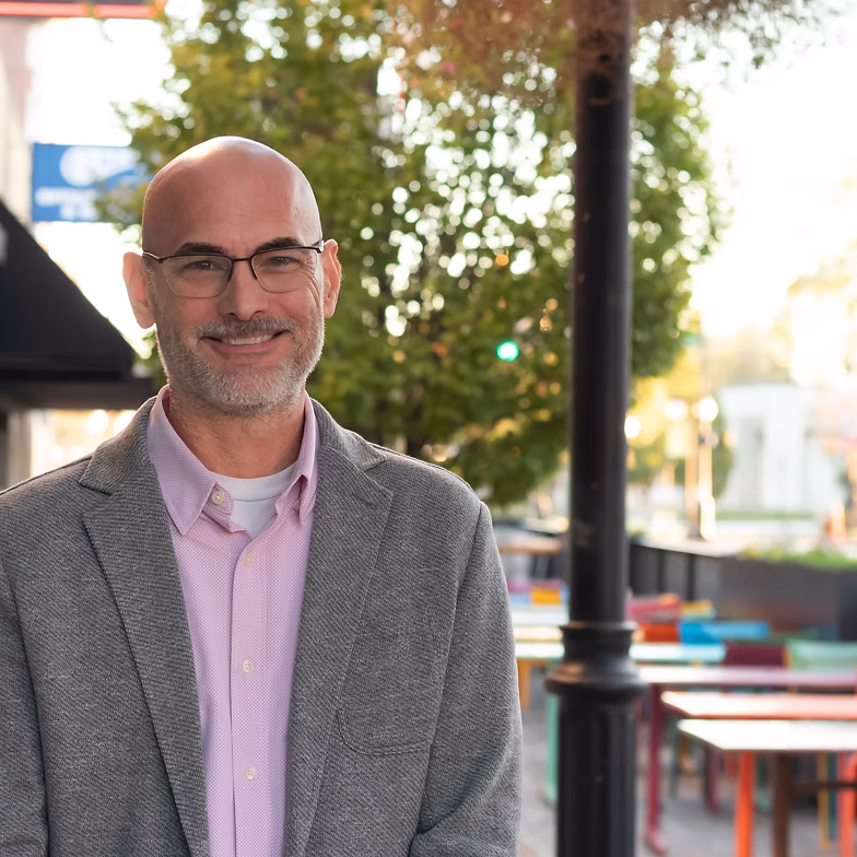 Neal Harwell with glasses and a beard wears a gray blazer and light purple shirt, standing outdoors by colorful tables and trees on a sunny day.