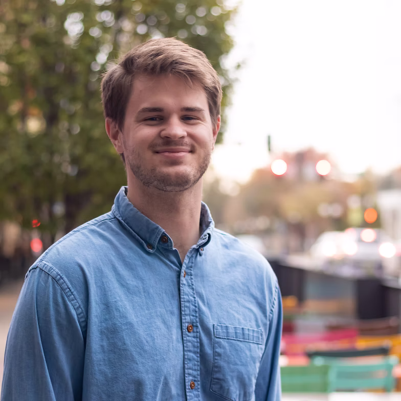 Cody Cottrell with short brown hair and a beard, wearing a light blue button-up shirt, smiles while standing outdoors on a city street with blurred trees and colorful benches in the background.