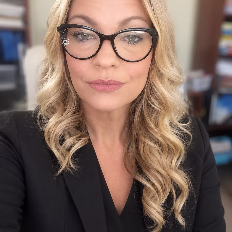 Amy Yang with long, wavy blonde hair and black-framed glasses wears a black blazer, looking directly at the camera. Shelves with books are blurred in the background.