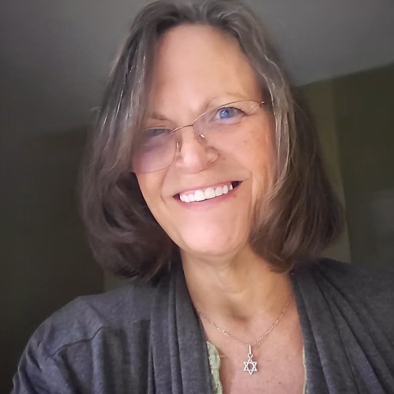 Cherryl Wardwell with shoulder-length brown hair, wearing glasses, a gray sweater, and a Star of David necklace, standing indoors with a softly lit background.