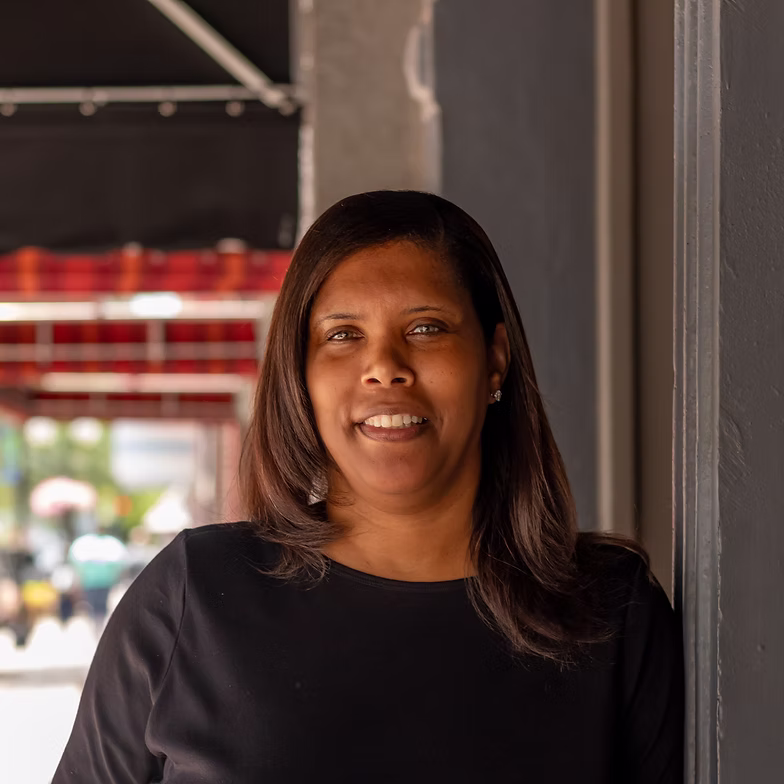 Group discussion-bro Laquintha Henderson with straight dark hair, wearing a black shirt, stands and smiles outdoors near a gray wall, with a blurred urban background behind her.