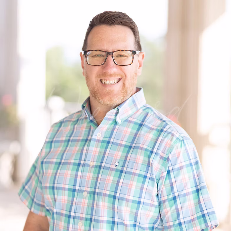 Patrick Luse with short hair and glasses wears a pastel plaid button-up shirt, standing outdoors in a brightly lit area with blurred columns and greenery in the background.