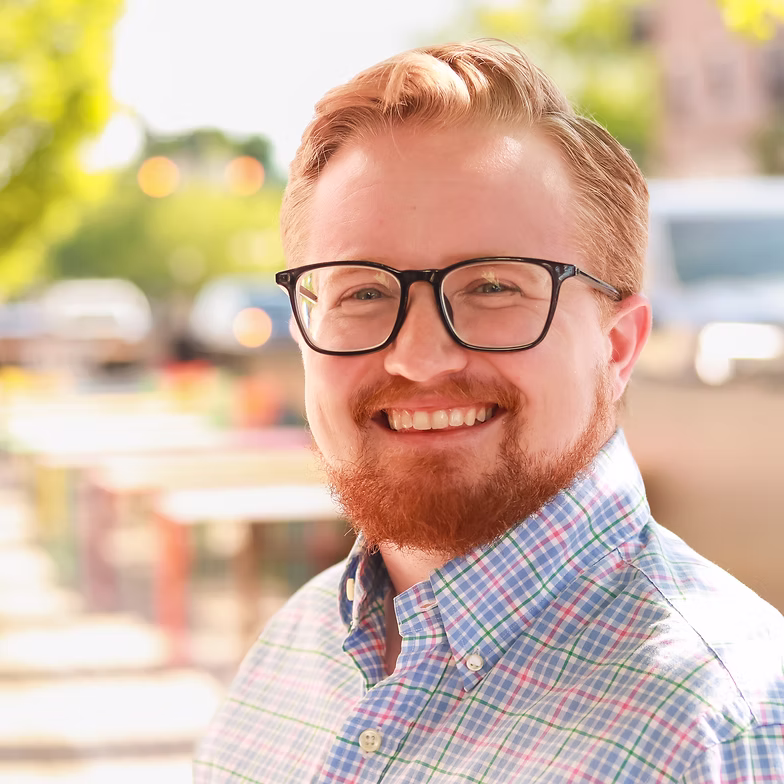 Group discussion-bro Austin Tisdale with short red hair, a beard, and glasses wears a plaid shirt and stands outdoors with blurred trees, lights, and tables in the background on a sunny day.