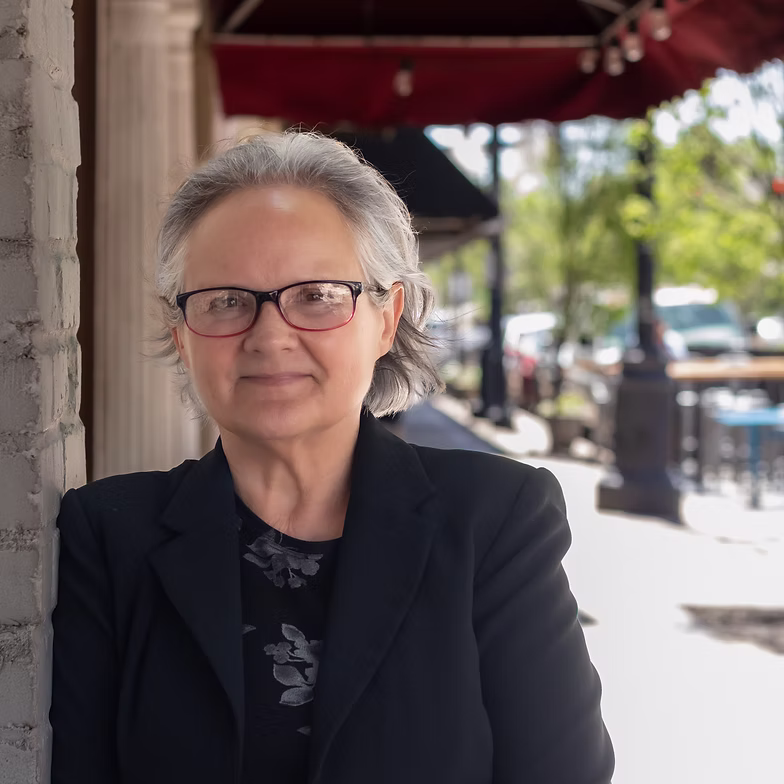 Sherry Clements with gray hair and glasses, wearing a black blazer, stands outside near a brick wall on a sunny day. The background shows a city street with trees, cars, and outdoor seating.
