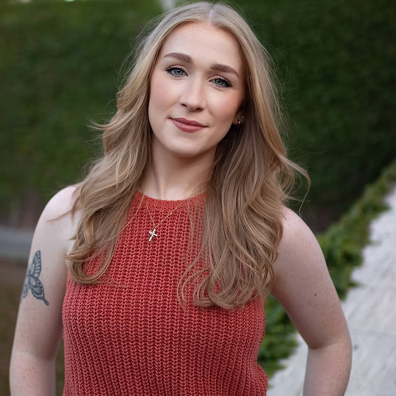 Kennedy Coker with long, wavy blonde hair wearing a sleeveless red knit top stands outside, smiling softly. She has a butterfly tattoo on her upper left arm and is wearing a necklace with a cross pendant.