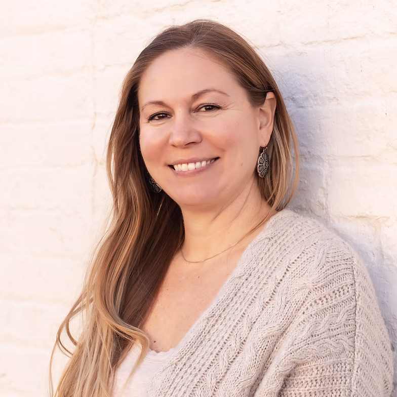Katherine Bishop with long light brown hair, wearing a beige sweater and earrings, smiles while standing against a white brick wall in soft natural light.