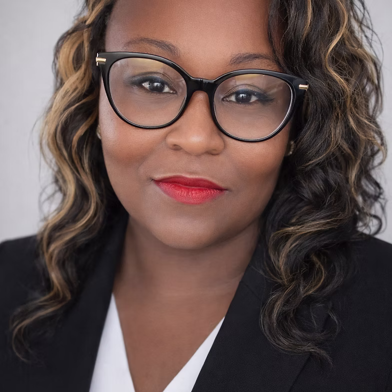 Group discussion-bro Sabrina Lewis with wavy hair, wearing black glasses, a black blazer, and red lipstick, looks confidently at the camera against a light background.