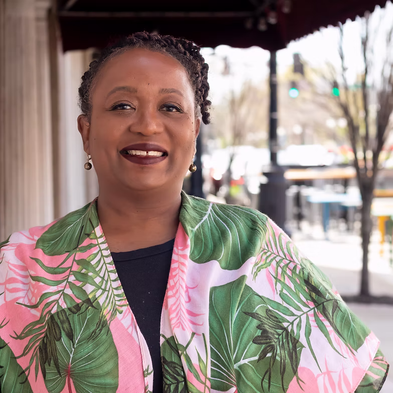 Fatimaah Carmichael-Anthony wearing a colorful, leafy-patterned jacket stands outdoors on a city sidewalk, with blurred buildings and trees in the background.