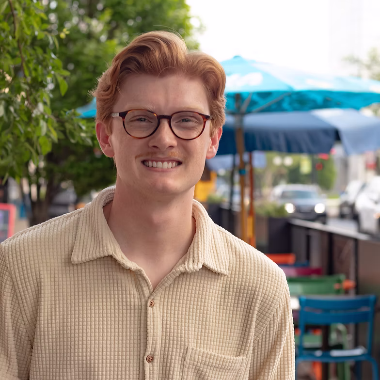 Carter Shields with red hair, glasses, and a beige shirt smiles outdoors. Colorful chairs and blue umbrellas are visible in the background on a sidewalk.