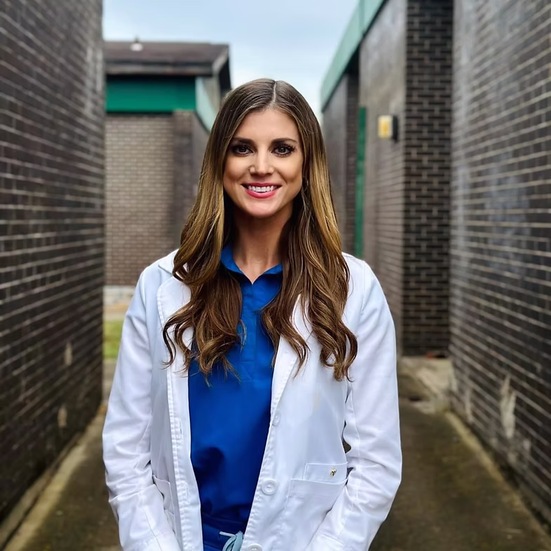 Melanie Higgins with long brown hair, wearing a blue shirt and white lab coat, stands and smiles in an outdoor alleyway with brick walls on both sides.