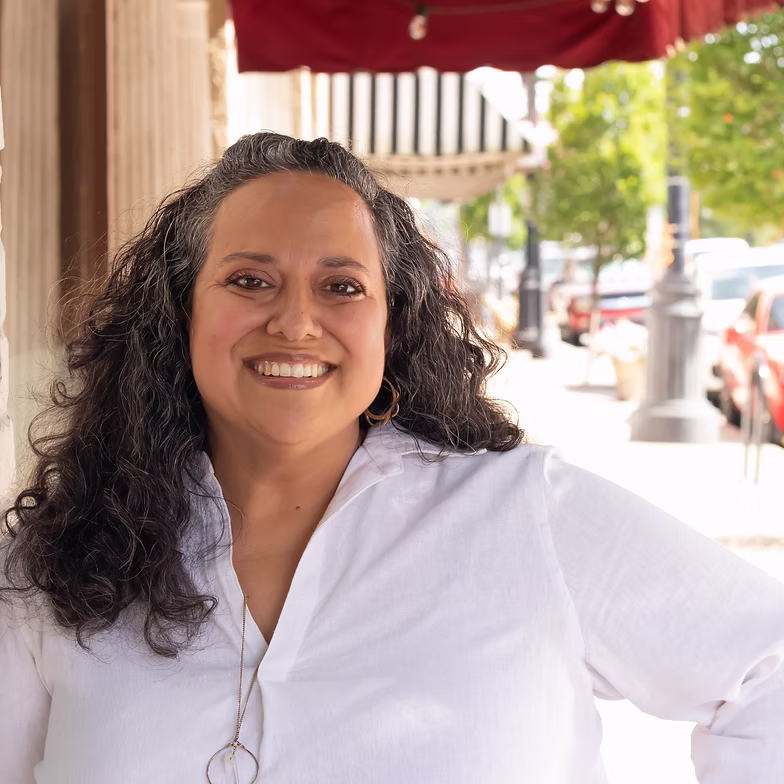Group discussion-bro Gabriela O’Dell with long curly hair wearing a white blouse stands outdoors on a city sidewalk, with trees, cars, and buildings in the background.