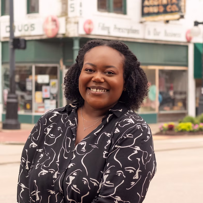 Group discussion-bro Demetra Thompson with curly hair stands on a sidewalk, wearing a black shirt with white abstract faces. A storefront and street signs are visible in the background.