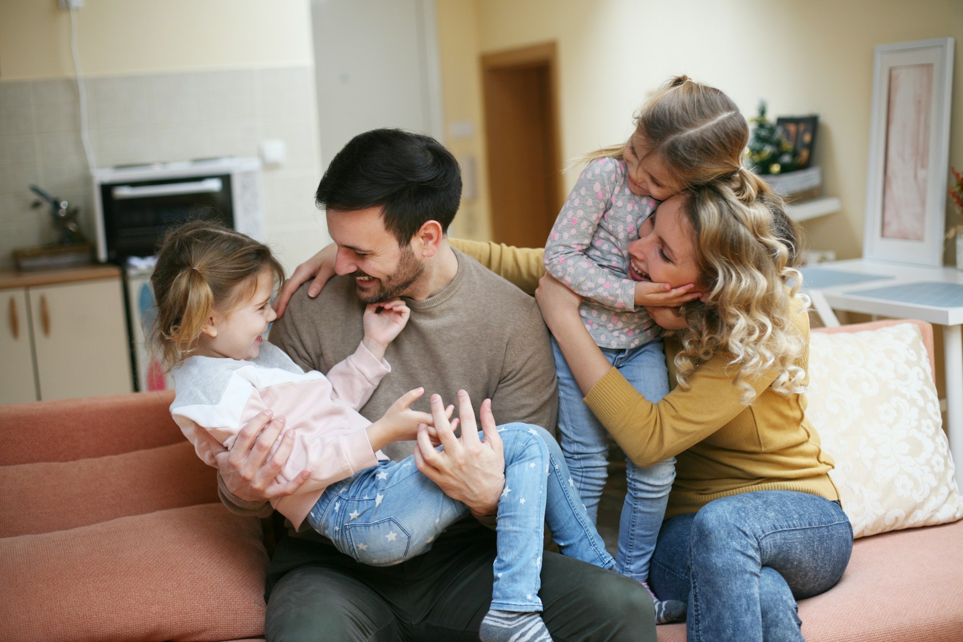 A photograph of a happy family of four sharing a joyful moment together on a living room couch. The parents are smiling warmly as the father cradles one young daughter in his lap while the mother holds and embraces their other daughter.